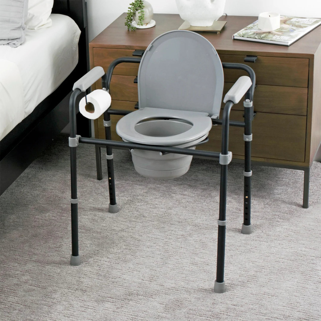 The Stander Universal Bedside Commode with TP Holder stands on a carpeted floor beside a wooden nightstand and bed, offering convenient bedside access with armrests and an integrated toilet paper holder.